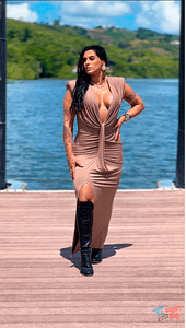 Modelo com vestido bege colado ao corpo e botas pretas, posando à beira d’água.