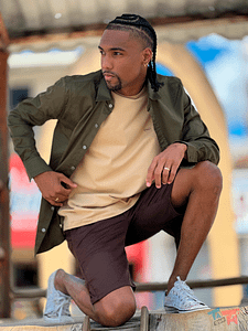 Homem posando com jaqueta verde musgo aberta, camiseta bege e bermuda marrom. Título: Camadas Masculinas para o Outono